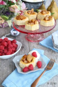 A serving of Pear Puff Pastry Tart on a dish with fresh red raspberries, setting in front of a cake stand full of puff pastry desserts.