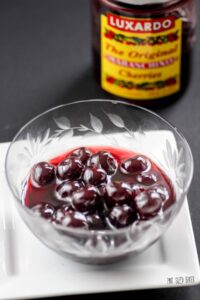 a glass bowl of Luxardo Cherries with the jar in the background.