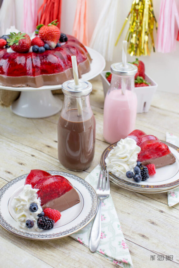 Slices of this stunning strawberry and chocolate jello mold served with fresh whipped cream and mixed berries along with chocolate milk and strawberry milk in small glass bottles.