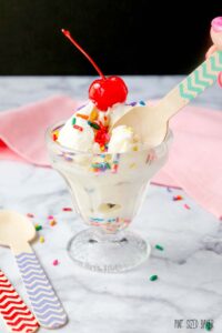 A scoop of vanilla ice cream with sprinkles and a cherry on top with a wooden spoon