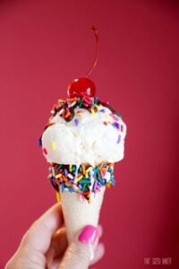 A hand holding a vanilla ice cream cone with chocolate, sprinkles and a cherry on top