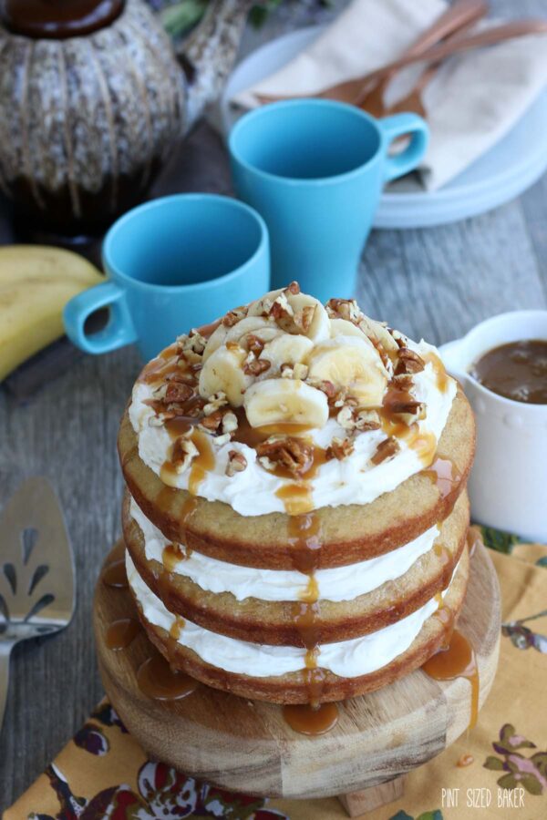 Banana naked cake recipe is finished and presented on a cake stand with coffee cups in the background. 