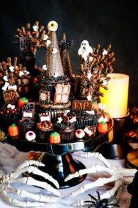 Chocolate cupcakes decorated like a haunted house on a black cake platter with skeleton hands at the bottom.