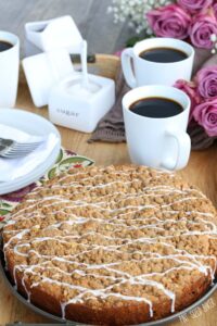 A rum raisin coffee cake with white icing drizzled on top sitting on a wooden cutting board in front of three cups of coffee.
