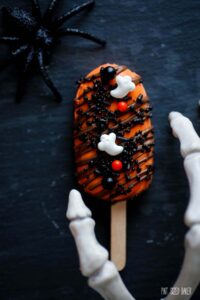 An orange cakesickle with black sprinkles and ghost candies.
