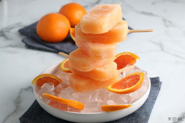 A horizontal picture of six Orange juice and Campari Popsicles stacked up on a bed of ice cubes and orange slices.