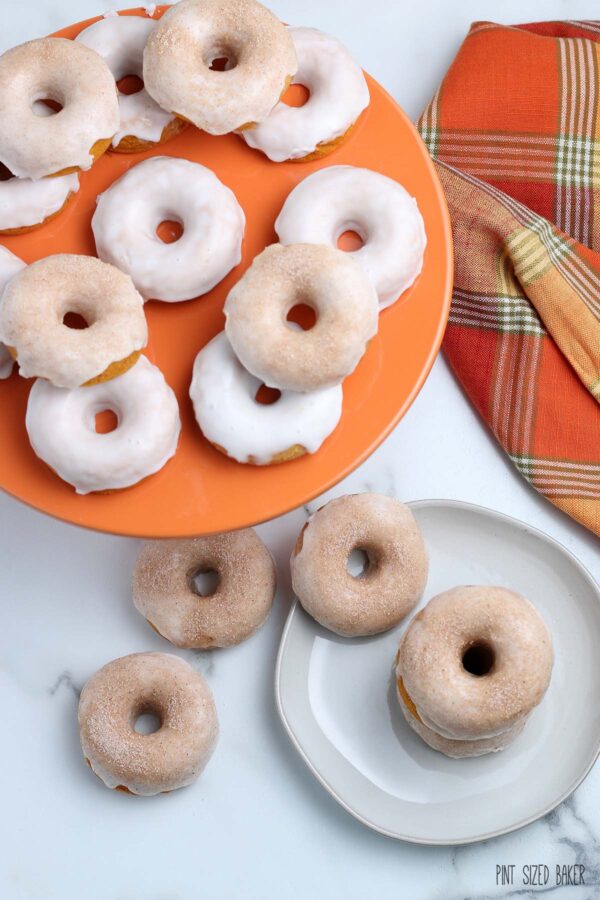 More baked pumpkin doughnuts piled on an orange cake stand and plate.