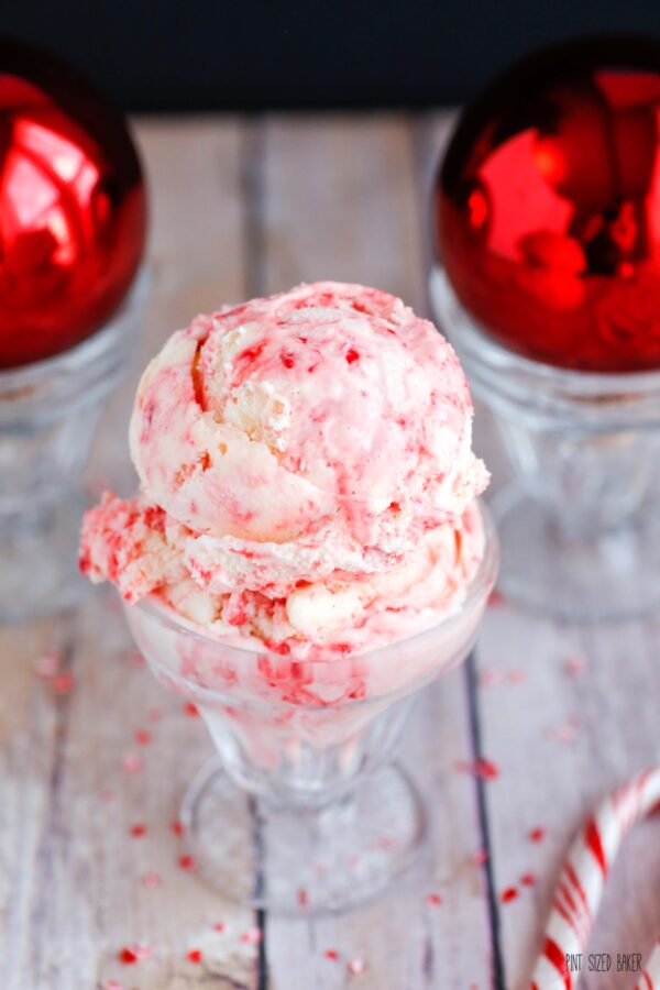 An image of two scoops of candy cane ice cream in a glass cup with bright red ornaments in the background.