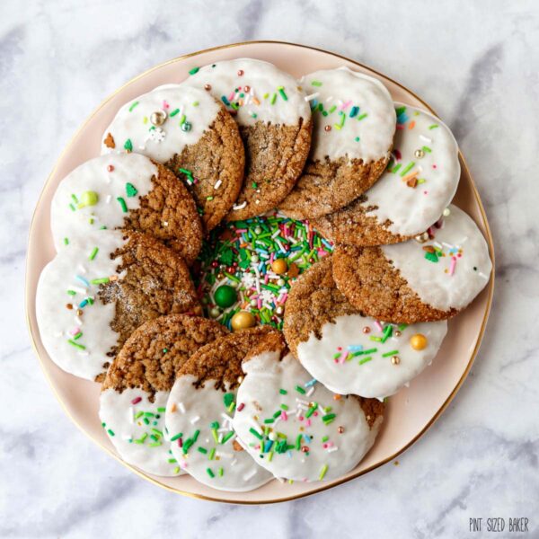 A square image of the molasses cookies dipped in white icing with green sprinkles in a circle on a pink plate.