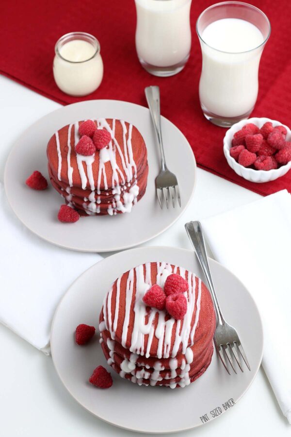 An image of two white plates stacked high each with a pile of pancakes on top with fresh raspberries.