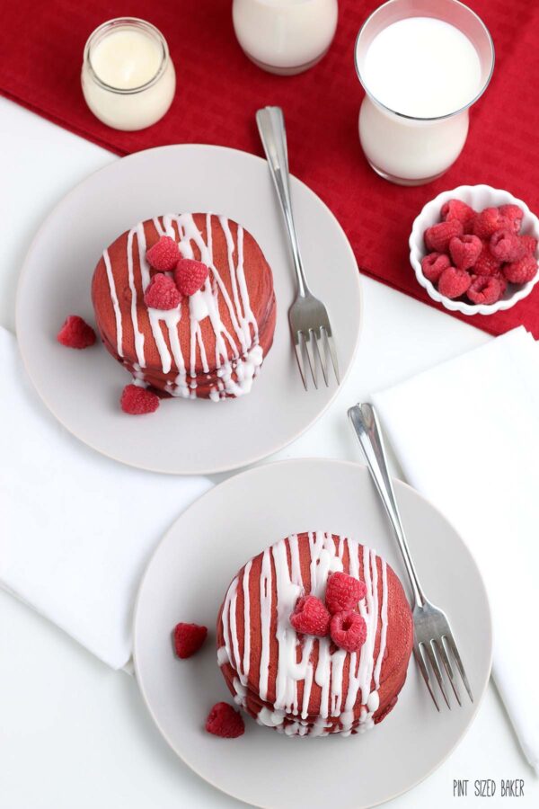 An overhead image of the red pancakes on while plates and white napkins with a red napkin in the background and a glass of milk.