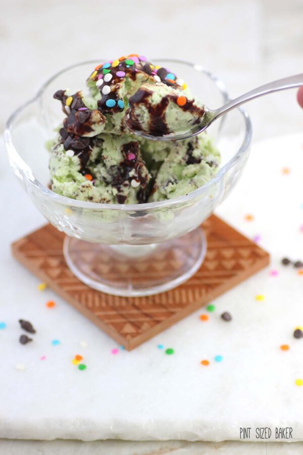 Green mint chip ice cream in a glass bowl with rainbow sequins on top and chocolate syrup.