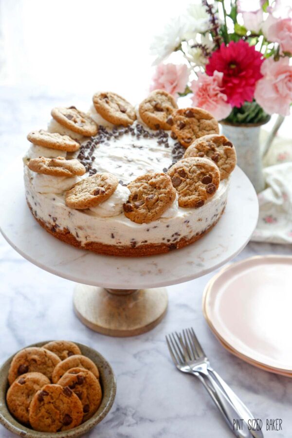 A no bake cheesecake with chocolate chip cookies on a marble cake pedestal with pink flowers in the background. 