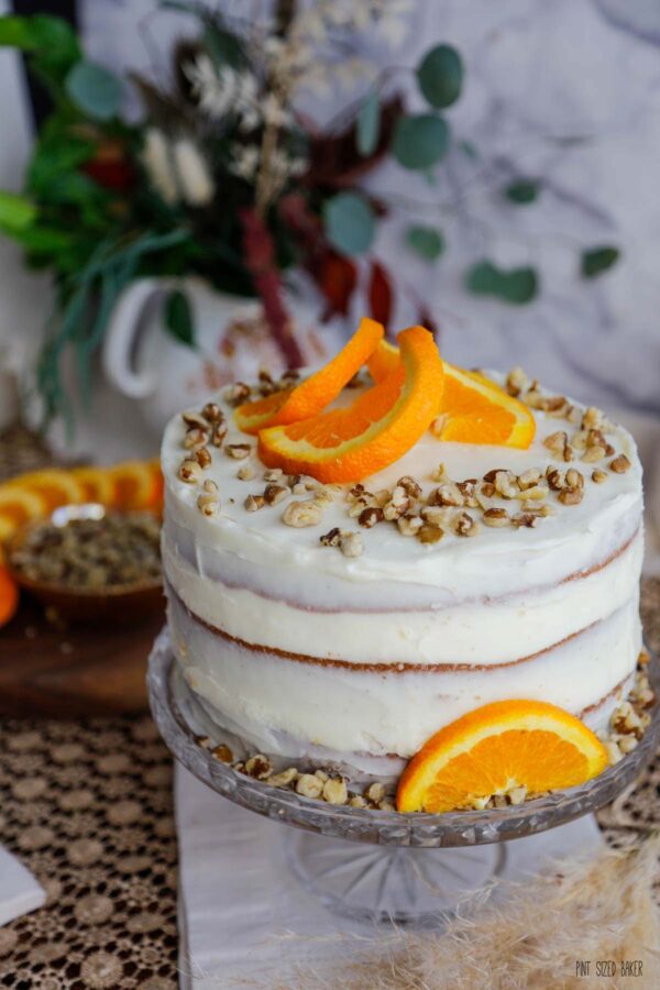 A photo of the finished cake showing the rough cake edges peaking though the cream cheese frosting with walnuts and orange slices on top.