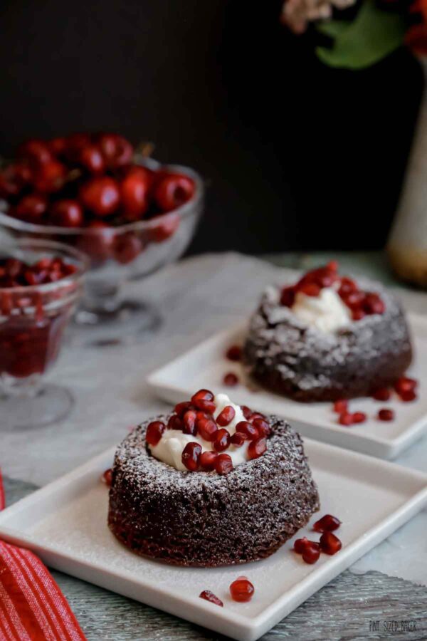 Two fudgy chocolate cakes on white plates served with whipped cream and pomegranate seeds.