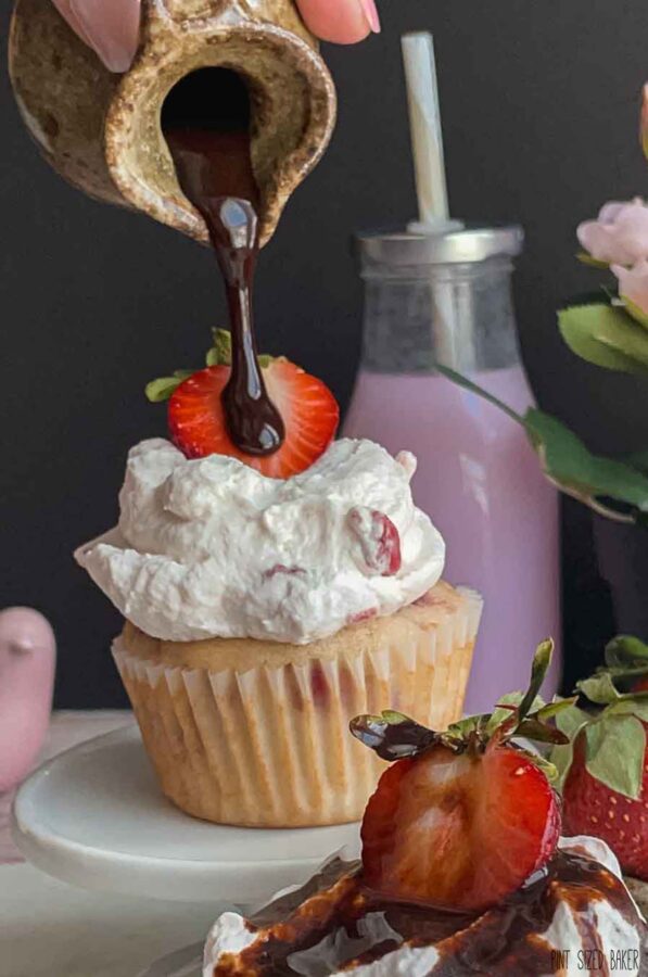 Strawberry Cupcake with strawberry whipped cream frosting a strawberry slice and chocolate syrup being drizzled over top.