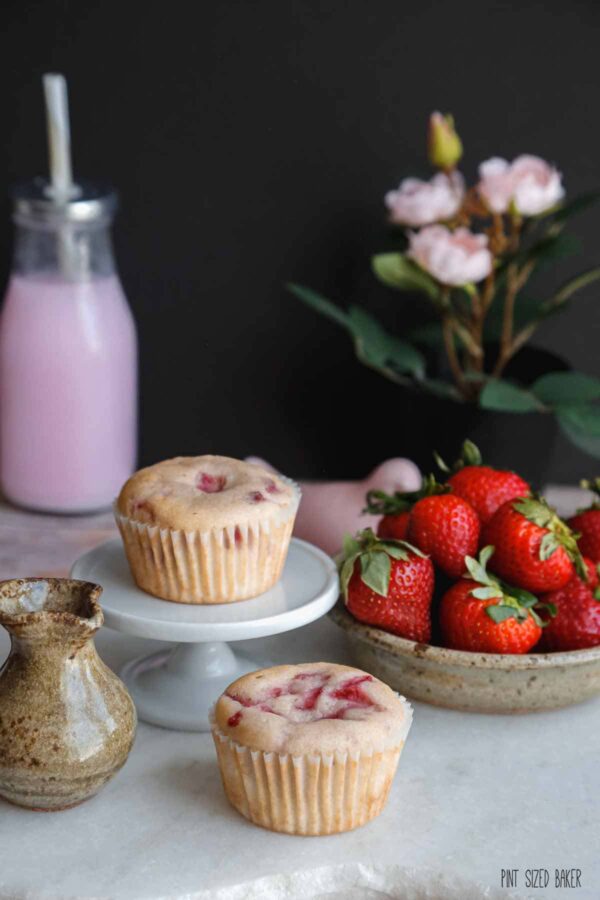 Plain strawberry cupcakes with a bowl of strawberries and a milk jug glass container with strawberry milk. 