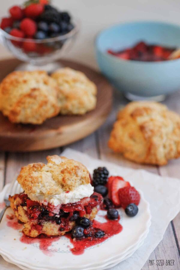 A shortcake full of mixed berries and fresh whipped cream on a plate with fresh baked biscuits in the background.