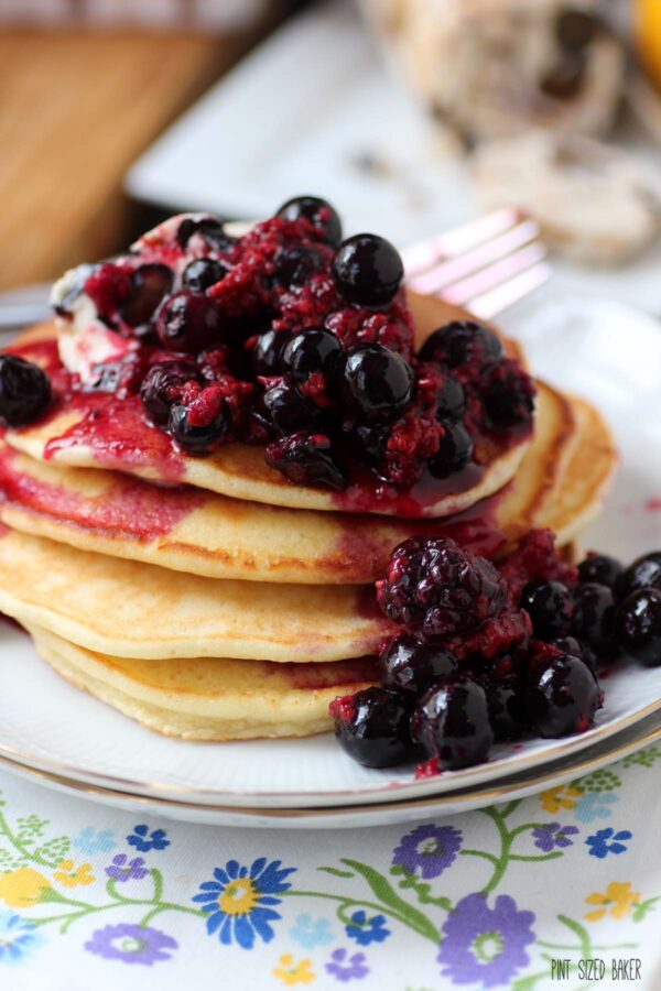 An close look at a stacked plate of lemon pancakes topped with loads of cooked blueberries, raspberries, and blackberries.