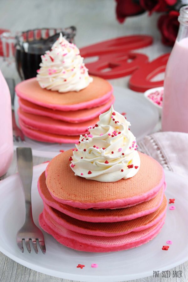 A giant stack of pancakes on a white plate and topped with a swirl of shipped cream and Valentine sprinkles.